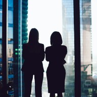 Back view of female colleagues in formal wear standing near window looking at modern exterior of skyscrapers in business center, silhouette of women together planning future success of brainstorming