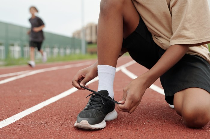 Hands of schoolboy tying black shoelace on gumshoe while standing on squats on race track against his classmates running along stadium