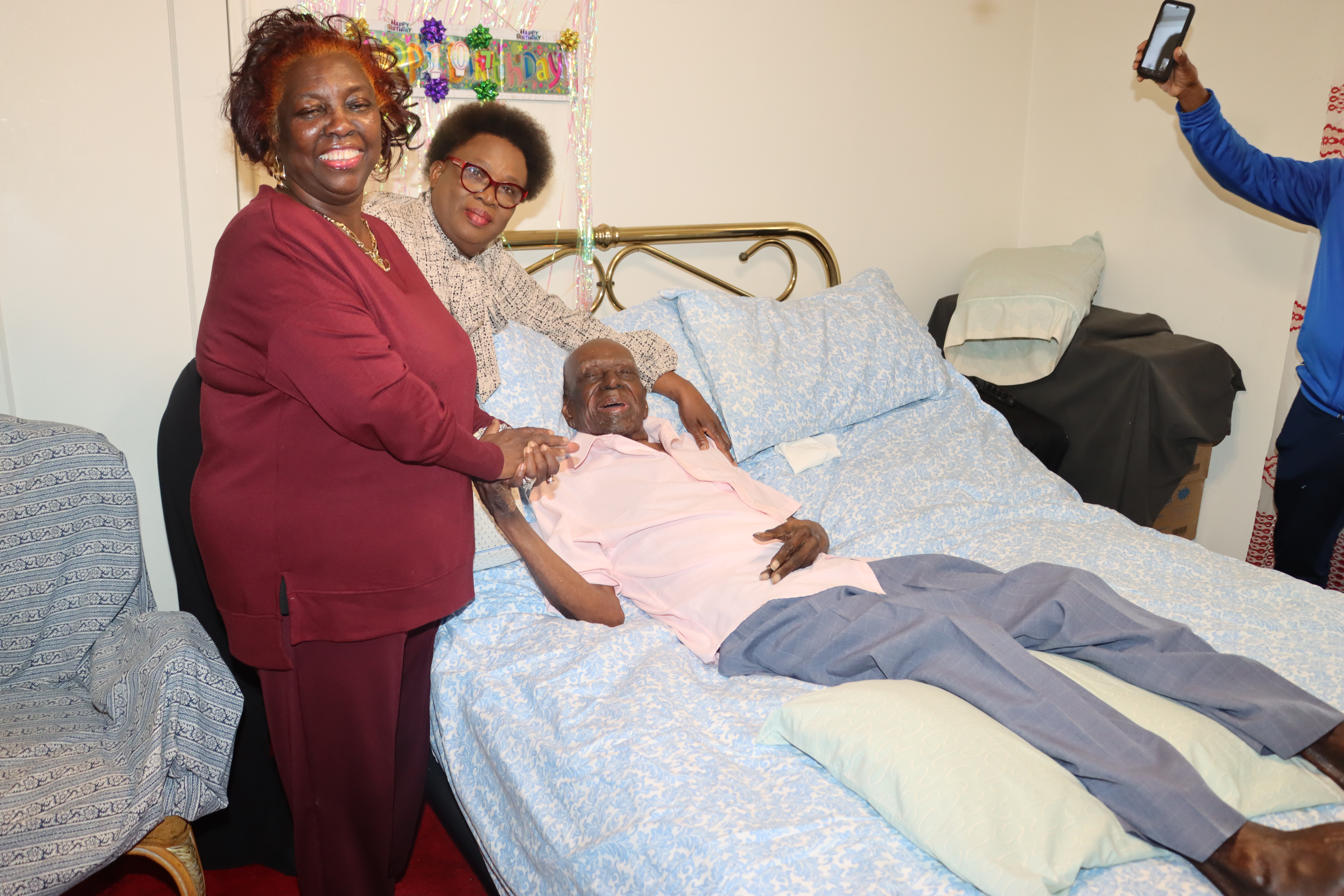 Ex-SVG Police Officers Cheryl Agard, foreground, and Pamella Ferrari-Easter celebrate Mitchinson "Mitchie" James's 107th birthday as he lays in bed